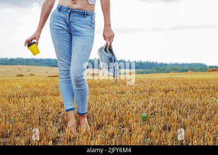 Barfuß Mädchen mit Turnschuhen und Pappbecher mit Kaffee in der Hand stehend auf dem landwirtschaftlichen Feld mit Heuhaufen und Ballen nach der Ernte. Stockfoto