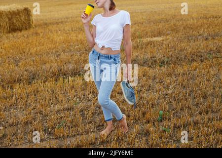 Barfuß Mädchen mit Turnschuhen und Pappbecher mit Kaffee in der Hand stehend auf dem landwirtschaftlichen Feld mit Heuhaufen und Ballen nach der Ernte. Stockfoto