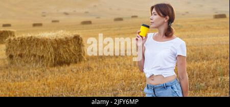 Barfuß Mädchen mit Turnschuhen und Pappbecher mit Kaffee in der Hand stehend auf dem landwirtschaftlichen Feld mit Heuhaufen und Ballen nach der Ernte. Stockfoto