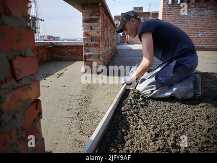 Seitenansicht eines jungen Arbeiters in Schutzhandschuhen, der die Betonplatte mit einer Kelle an der frischen Luft ausebelt. Konzept der Prozessarbeit mit Zement und speziellen Ausrüstungen bei gutem Wetter. Stockfoto