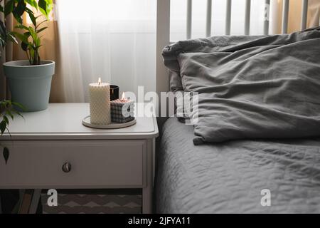 Schlafzimmer mit heller Innenausstattung. Moderne Kerze, Hauspflanze auf Nachttisch neben dem Bett. Nach Hause duftendes Aroma. Wellness Stockfoto