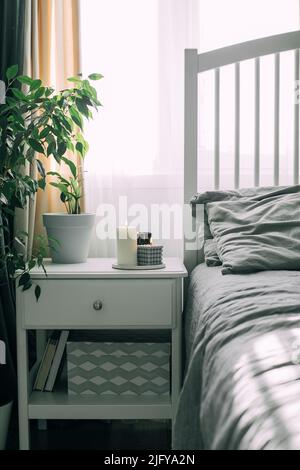 Schlafzimmer mit heller Innenausstattung. Moderne Kerze, Hauspflanze auf Nachttisch neben dem Bett. Nach Hause duftendes Aroma. Wellness Stockfoto