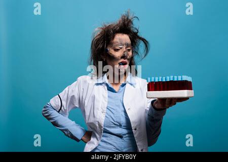 Mad Biochemie Spezialist mit schmutzigem Gesicht und unordentliche Frisur halten Tablett mit Glas-Reagenzgläser, während auf blauem Hintergrund. Verrückter Wissenschaftler mit einem Reagenzglas zwischen den Zähnen, das unvorsichtig auf Glaswaren schaut. Stockfoto