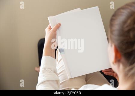 Schöne weibliche Hände halten ein offenes Buch oder eine Zeitschrift in einem Raum auf einem schwarzen Sofa Stockfoto