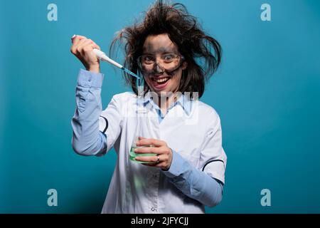 Verrückter Wissenschaftler grinsend gruselig, während er chemische Verbindungen von der Pipette in einen mit flüssiger Substanz gefüllten Glaskolben tropfte. Verrückter, törichter Laborarbeiter, der Pipette verwendet, um experimentelles Serum zu mischen. Studioaufnahme Stockfoto