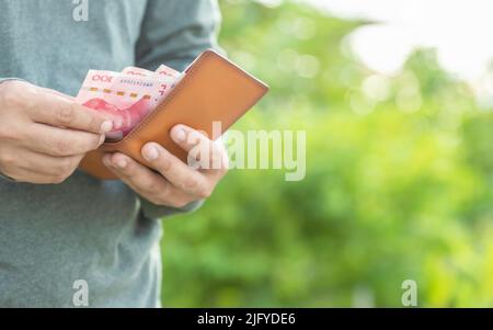 Asiatischer Mann zählt und überprüft chinesische Banknoten in seiner Brieftasche. Außenaufnahmen mit grün verwacklungsunscharfer Hintergrundfarbe Stockfoto