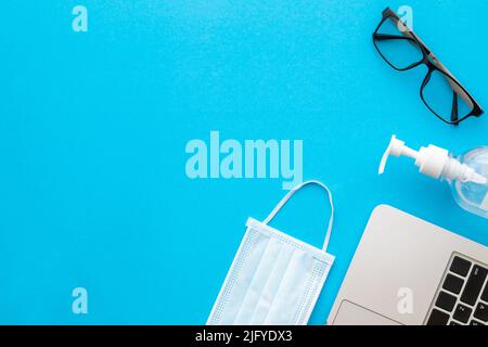 Draufsicht neue präventive Maske, Desinfektionsmittel-Gel, Laptop und Augenbrille auf blauem Hintergrund mit Platz für Text oder Design. Coronavirus-Krankheit (COVID- Stockfoto