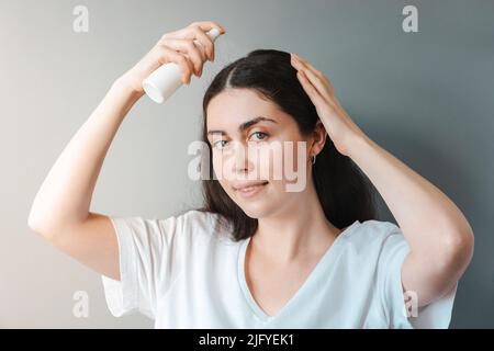 Eine junge schöne Frau sprüht Pflegekosmetik auf ihr Haar. Grauer Hintergrund. Das Konzept der Haarpflege. Stockfoto