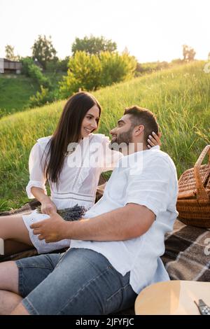 Brünette Frau in weißem Kleid lächelnd auf bärtigen Mann auf Picknick auf dem Land Stockfoto