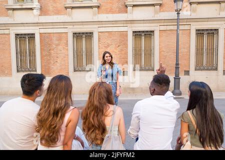 Mann, der einem Reiseleiter vor einer Gruppe von Freunden auf einem öffentlichen Platz eine Frage stellt Stockfoto