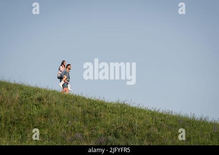 Blick aus der Ferne auf Mann Huckepack Freundin auf grasbewachsenen Hang Stockfoto