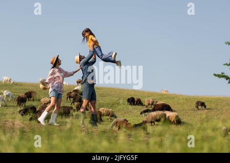 Seitenansicht des aufgeregt Landwirts, der seine Tochter in der Nähe von Frau und Schafherde auf einer grünen Wiese hält Stockfoto