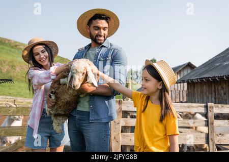 Frau und Kind in Strohhüten streichelten Lamm in den Händen eines lächelnden Bauern Stockfoto