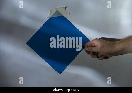 Die Hand eines reifen Mannes zeigt mit einem Bleistift auf ein Stück Papier. Leeres blaues quadratisches Stück Papier, das an der Wand befestigt ist. Eine abstrakte Licht- und Schattenzeichnung o Stockfoto