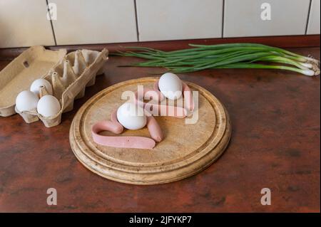 Zwei herzförmige Würstchen liegen auf einem Schneidebrett. Ein Tablett mit Hühnereiern und einem Bund grüner Zwiebeln stehen nebeneinander. Kochen der Mahlzeit. Liebe, Stockfoto