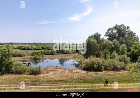 TSCHERNIHIW REG, UKRAINE - 19. Juni 2022: Krieg in der Ukraine. Ein einsamer Mann geht mit einem ausgebrannten Bus und einem zerstörten Auto auf einem Weg in der Nähe eines Sees um sein Geschäft Stockfoto