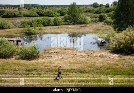 TSCHERNIHIW REG, UKRAINE - 19. Juni 2022: Krieg in der Ukraine. Ein einsamer Mann geht mit einem ausgebrannten Bus und einem zerstörten Auto auf einem Weg in der Nähe eines Sees um sein Geschäft Stockfoto