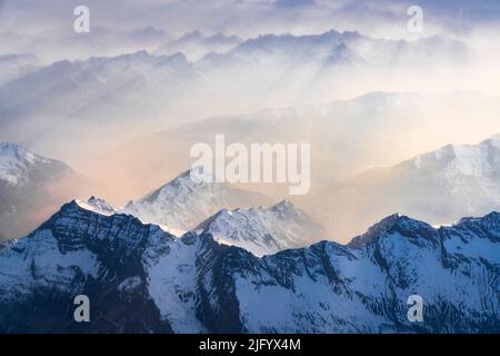 Sonnenstrahlen durch Nebel über den schneebedeckten Gipfeln der Lepontine und der Tessiner Alpen bei Sonnenuntergang vom Flugzeug aus, Schweiz, Europa Stockfoto