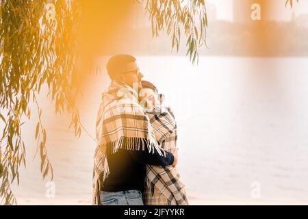 Verliebtes Paar, das sich im kalten goldenen Herbst unter gelben Blättern umarmt und mit einer warmen Decke auf dem Strandwasser bedeckt. Ein Tag im Park. Defokussieren Sie den Vordergrund. Stockfoto