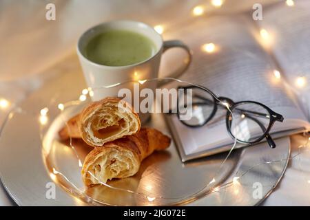 Croissants, Matcha-Tee, Buch und Gläser im Bett Stockfoto