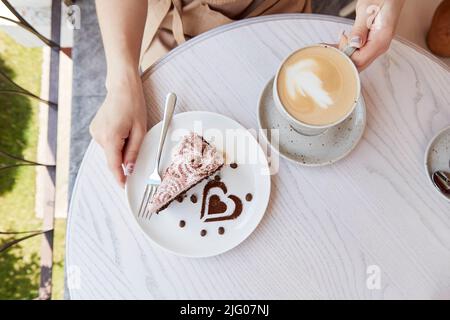 Frau Hände halten Obst Schokolade Tasse Cappuccino und Kuchen auf Holztisch in der Café-Terrasse draußen. Atmosphärische Kaffeezeit. Dessertdekorationen in Herzform Stockfoto