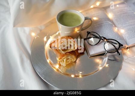 Croissants, Matcha-Tee, Buch und Gläser im Bett Stockfoto