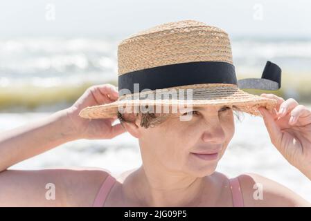 Grauhaarige Frau mit einem leichten Lächeln, das die Kanten ihres Hutes stützt, die sich vom Meer winden und versuchen, ihren Kopf abzureißen und dabei etwas am Ufer zu beobachten. Stockfoto