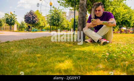 Ein junger Mann mit Dreadlocks ruht im Schatten eines Baumes und nutzt ein Smartphone im Park. Ein gutaussehender Kerl mit Sonnenbrille sitzt auf dem Gras Stockfoto