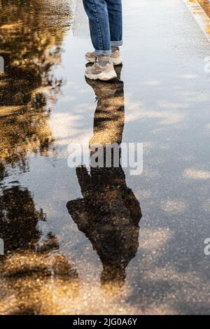 Beine der Frau in Jeans und Sneakers. Die Silhouette einer Frau spiegelt sich in einer Pfütze auf dem Bürgersteig wider. Lauf dem Regen nach. Stockfoto