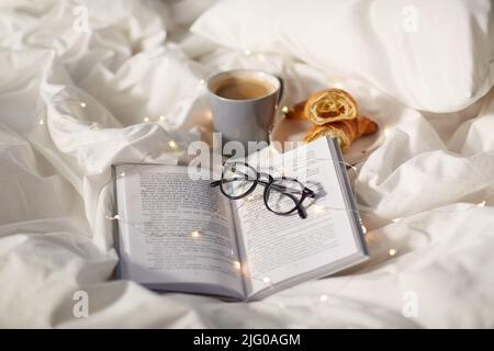 Croissants, Tasse Kaffee, Buch und Gläser im Bett Stockfoto