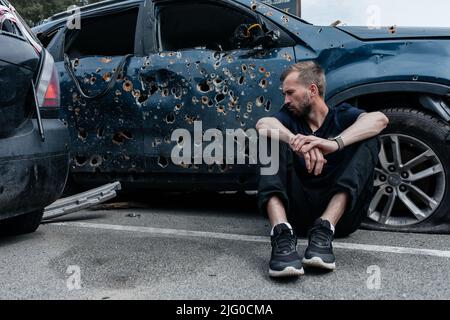 Mann sitzt in der Nähe von Schuss beschädigt Auto gespickt mit Kugeln. Auto, das während der Evakuierung von Zivilisten vom russischen Militär angeschossen wurde. Schrapnel und Einschusslöcher Stockfoto