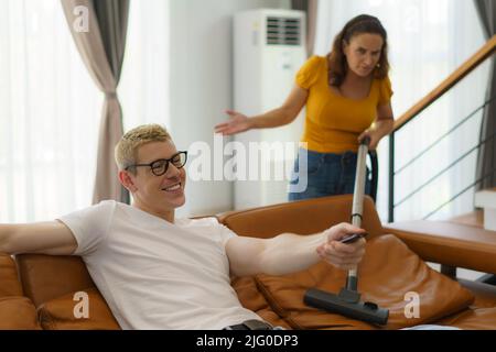 Die kaukasische Frau, die im Wohnzimmer des Hauses einen Staubsauger benutzte, verärgerte ihren Mann, weil er bei der Hausarbeit nicht mitgeholfen hatte, und setzte sich bequem hin und gucken fern. Stockfoto