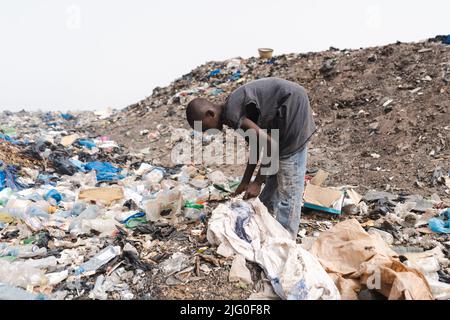 Junger afrikanischer Müllsammler, der in einer städtischen Deponie einen Plastikbeutel mit wiederverwendbaren Gegenständen füllt; informelles Abfallrecycling-Konzept Stockfoto