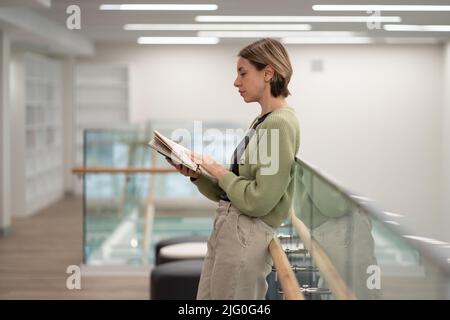 Nachdenkliche skandinavische Frau mittleren Alters, die in der Bibliothek Interessantes Buch liest Stockfoto