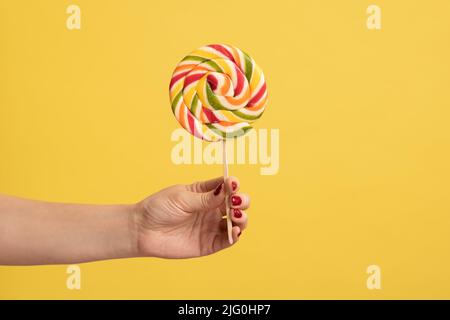 Nahaufnahme der Frau Hand hält appetitliche Runde Regenbogenbonbon auf Stick, große Lollipop in Arm, Süßwaren Werbung, Glukose süße Lebensmittel. Innenaufnahme des Studios isoliert auf gelbem Hintergrund. Stockfoto