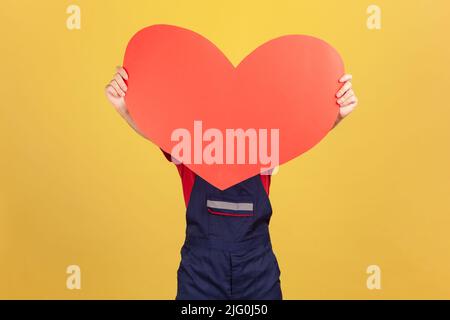 Porträt eines unbekannten anonymen Arbeiters, der ein großes rotes Herz in der Hand hält und sein Gesicht hinter einem papierromantischen Symbol versteckt, Lieferung am Valentinstag. Innenaufnahme des Studios isoliert auf gelbem Hintergrund. Stockfoto