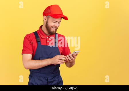 Porträt eines zufriedenen lächelnden Kuriers oder Handwerkers, der mit dem Smartphone in den Händen steht, die Online-Bestellung übernimmt und bereit zur Arbeit ist. Innenaufnahme des Studios isoliert auf gelbem Hintergrund. Stockfoto