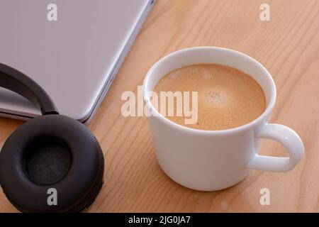 Eine Nahaufnahme einer Tasse Kaffee mit Kopfhörer und Laptop auf der Seite auf einem Holztisch Stockfoto