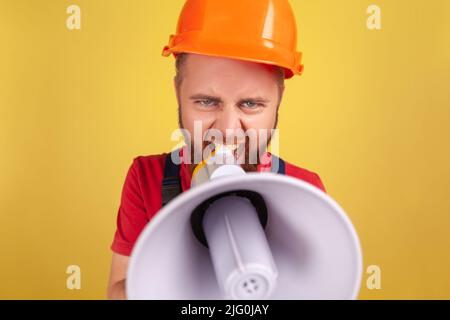 Porträt eines wütenden, aggressiven Arbeiters, der Uniform und Schutzhelm trägt und Selfie macht, aus der Perspektive eines Fotos, schreiend im Megaphon. Innenaufnahme des Studios isoliert auf gelbem Hintergrund. Stockfoto