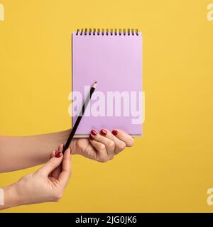 Seitenansicht Nahaufnahme einer Frau, die einen violetten Notizblock in der Hand hält und leeres Papier mit Stift oder Bleistift zeigt. Innenaufnahme des Studios isoliert auf gelbem Hintergrund. Stockfoto