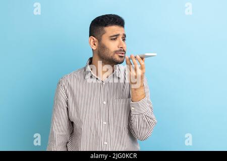 Porträt eines selbstbewussten jungen Geschäftsmannes, der das Mobiltelefon in der Hand hält und spricht, Voicemail hinterlässt und ein gestreiftes Hemd trägt. Innenaufnahme des Studios isoliert auf blauem Hintergrund. Stockfoto