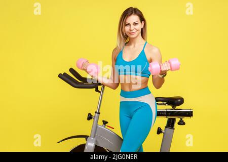 Porträt einer schönen blonden Frau Fitness-Ausbilderin mit Hanteln für Bizeps nach dem Cardio-Training, trägt blaue Sportkleidung. Innenaufnahme des Studios isoliert auf gelbem Hintergrund. Stockfoto