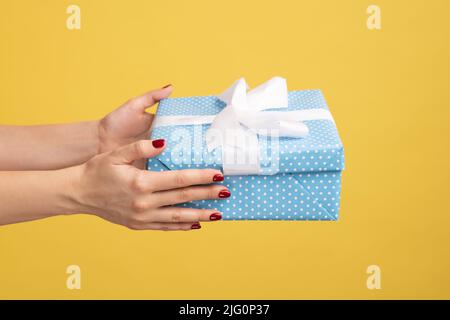 Nahaufnahme der Frau Hand hält verpackt blauen Geschenkkarton mit weißem Streifen, Lieferung des Geschenks an Feiertagen, Hand geben Geburtstagsüberraschung, Bonus. Innenaufnahme des Studios isoliert auf gelbem Hintergrund. Stockfoto