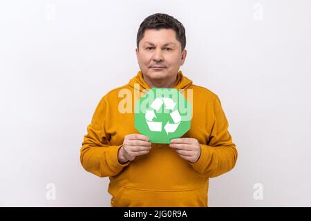 Positiv lächelnder Mann mit dunklem Haar und grünem Abfallrecycling-Symbol, zufrieden mit der Umweltsicherheit, trägt einen Hoodie im urbanen Stil. Innenaufnahme des Studios isoliert auf weißem Hintergrund. Stockfoto
