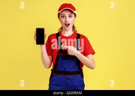 Porträt einer staunenden Arbeiterin, die mit einem Mobiltelefon mit leerem Display für Werbung steht, auf das Gadget zeigt, Overalls und rote Mütze trägt. Innenaufnahme des Studios isoliert auf gelbem Hintergrund. Stockfoto