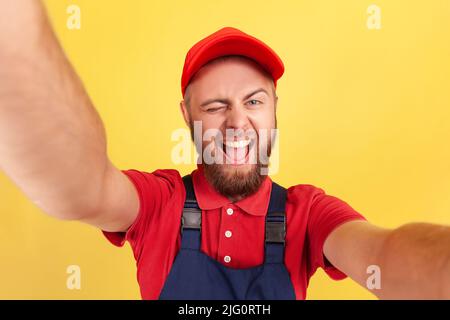 Porträt eines lustigen, verspielten bärtigen Arbeiters in Uniform, der Selfie macht, auf den Blickwinkel der Kamera schaut und winkelt, aus der Perspektive des Fotos. Innenaufnahme des Studios isoliert auf gelbem Hintergrund. Stockfoto