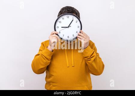 Porträt eines anonymen unbekannten Mannes, der sich hinter einer großen Wanduhr versteckt, Zeitmanagement, erinnert an Deadline, trägt einen Hoodie im urbanen Stil. Innenaufnahme des Studios isoliert auf weißem Hintergrund. Stockfoto