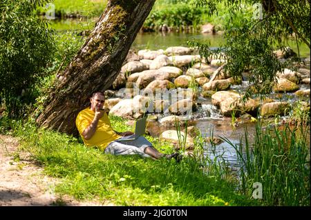 Mann mit Laptop und Smartphone, der draußen auf der Wiese am Fluss arbeitet, Open-Air-Bürokonzept Stockfoto
