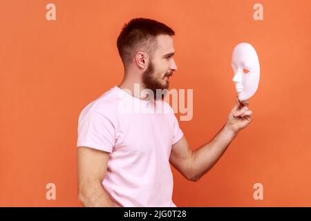 Seitenansicht Porträt eines bärtigen Mannes, der mit aufmerksamem Blick auf die weiße Maske in den Händen schaut und versucht, die verborgene Persönlichkeit zu verstehen, indem er ein rosa T-Shirt trägt. Innenaufnahme des Studios isoliert auf orangefarbenem Hintergrund. Stockfoto