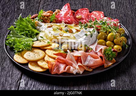 Herzhaft gebackener Brie mit Crackern, spanischem Salchichon, Salami-Würsten, dünn geschnittenem geräuchertem Schinken, grünen Oliven, Salat, Erdnüssen, Pecan auf schwarzer Platte Stockfoto